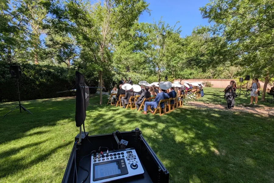 An outdoor ceremony under the trees at Los Poblanos, guests seated in the shade with Cutmaster Music sound equipment set up nearby